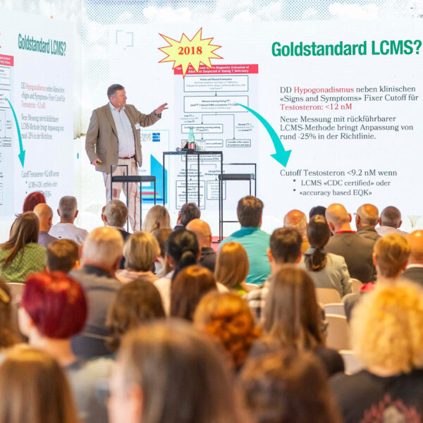 Mitten in seinem Vortrag: Christoph Seger steht auf der Bühne des Science Dialogue in Wien. Er steht zwischen zwei Leinwänden und weisst auf die Korrektur des Goldstandards hin.